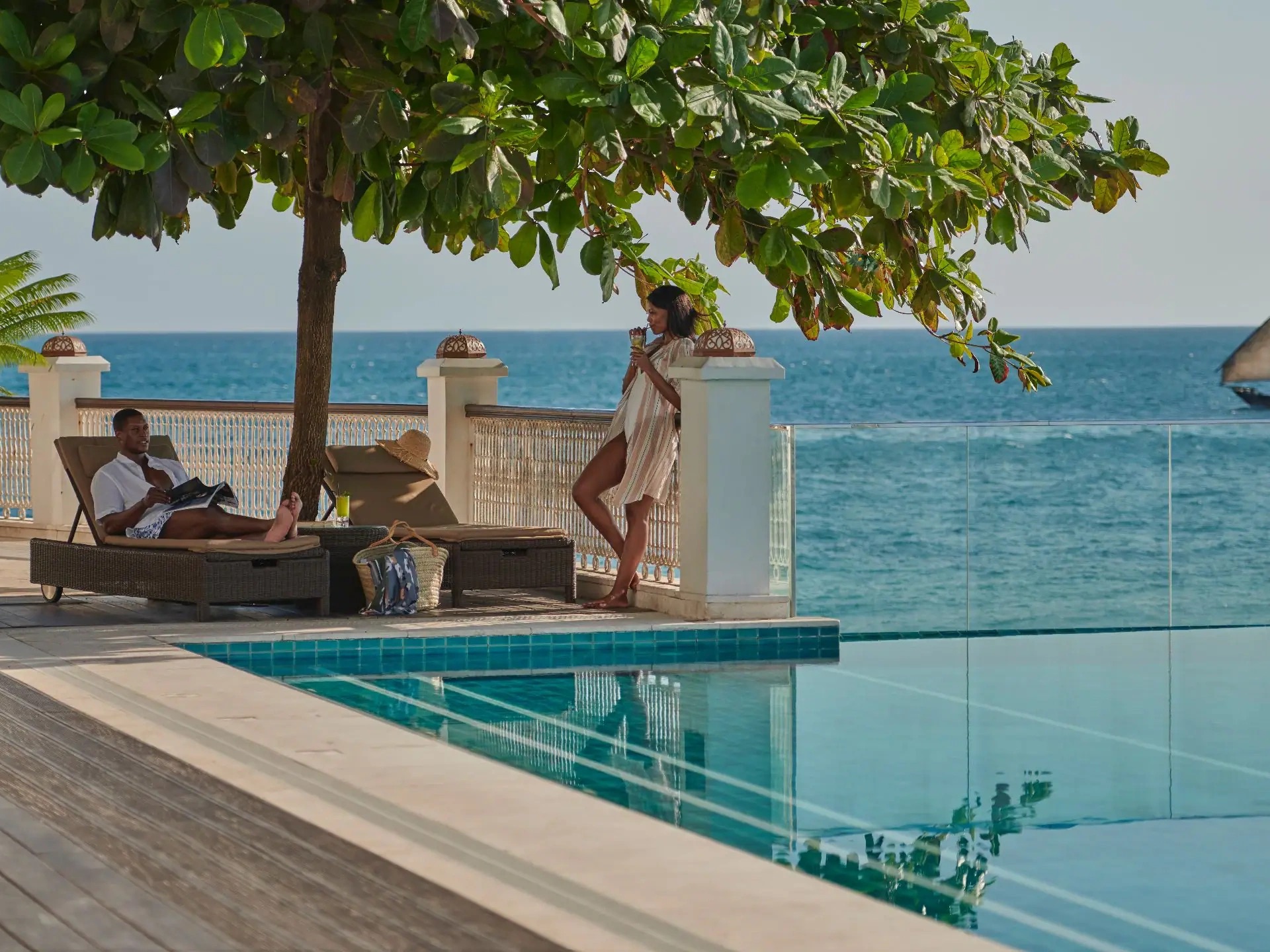 A couple beside an infinity pool on a seafront terrace, a dhow passing on the water beyond